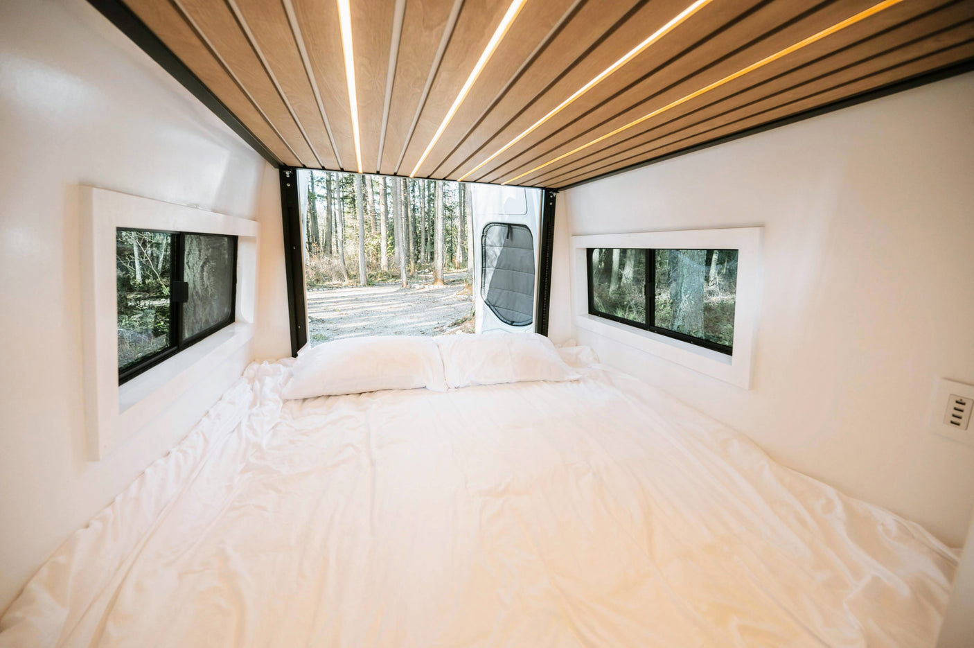 Bright RV rental camper van interior: white bed, large windows framing a forest, and modern wood-slat ceiling lights.
