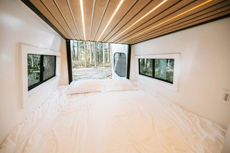 Bright RV rental camper van interior: white bed, large windows framing a forest, and modern wood-slat ceiling lights. — photo 11