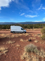 White campervan RV rental parked on a red dirt road, surrounded by desert brush and distant mountains under a blue sky.