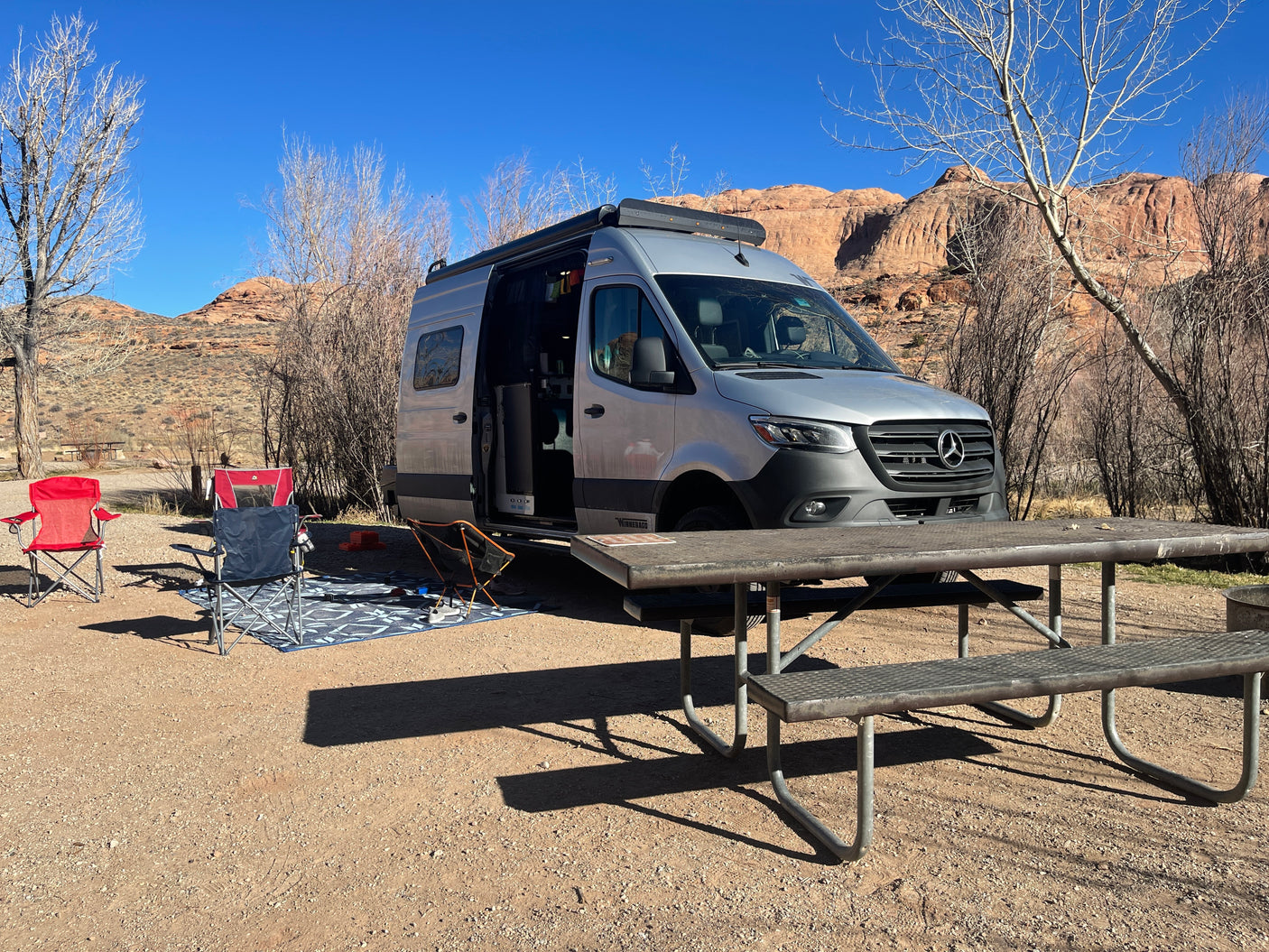 Scenic RV rental campsite: A silver camper van parked, with chairs, picnic table, and desert mountains.