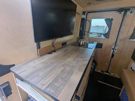 Modern RV rental kitchen counter with sink, induction cooktop, and mounted TV, built into a wooden interior.