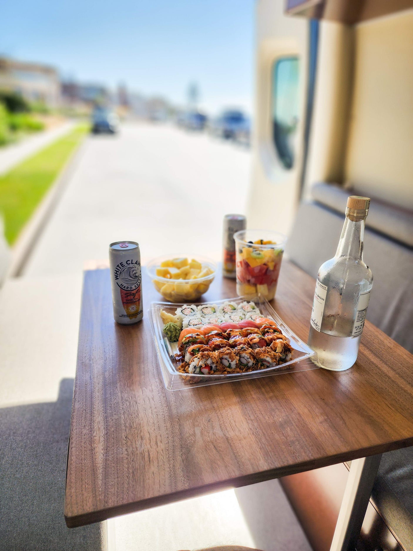 RV rental stop: a table laden with fresh sushi, fruit cups, White Claw, and a bottle of agave spirit, parked by a street.