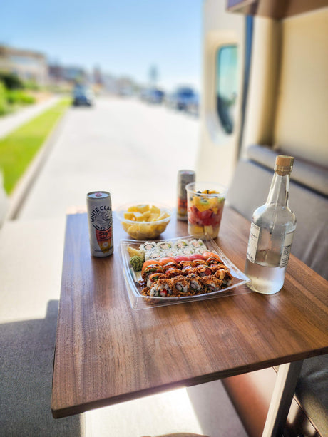 RV rental stop: a table laden with fresh sushi, fruit cups, White Claw, and a bottle of agave spirit, parked by a street.