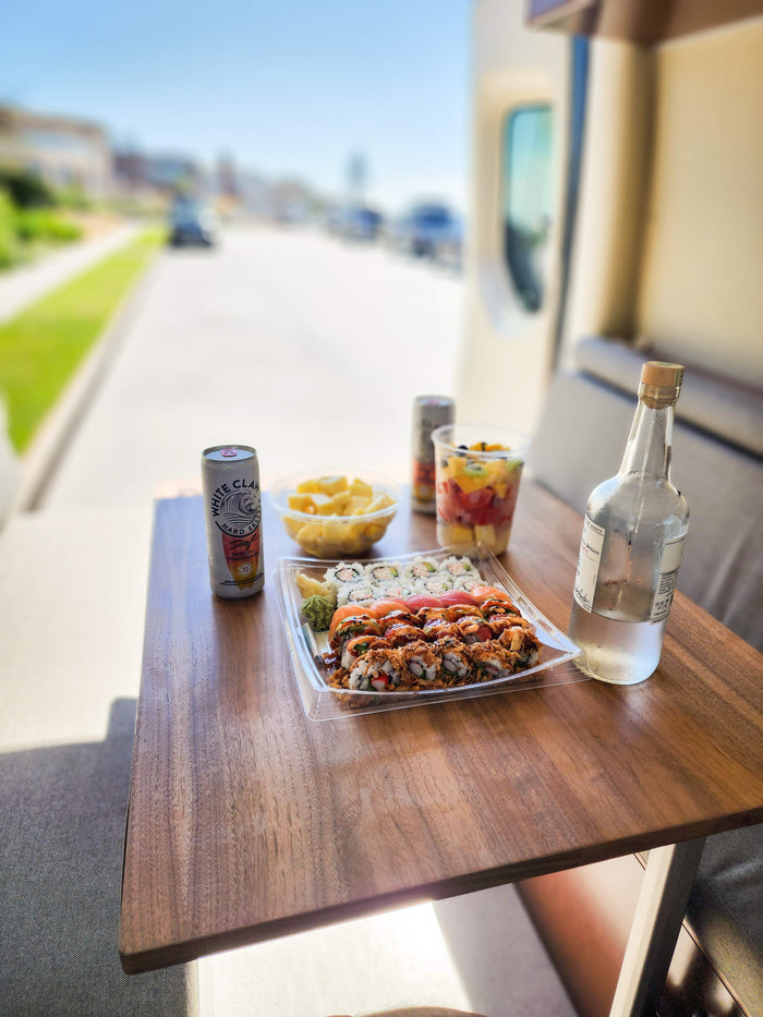 RV rental stop: a table laden with fresh sushi, fruit cups, White Claw, and a bottle of agave spirit, parked by a street.