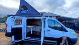A happy man in a blue shirt gives a thumbs-up from a pop-up rooftop tent on this white RV rental camper van, revealing its cozy interior.