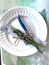 Perfect RV rental meal setup: plate, rosemary-tied napkin, blue knife on a nature-themed placemat. Enjoy glamping!