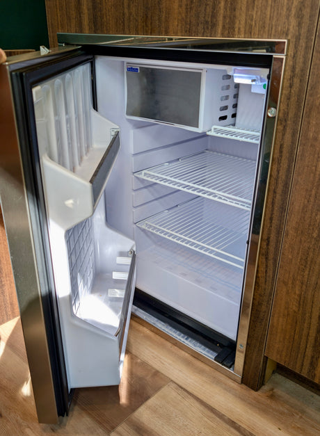 An open, empty mini-fridge inside an RV rental. Features include wire shelves, a small freezer, and integrated design within wooden cabinetry.