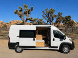 White **RV rental** camper van with open door revealing its interior, parked among Joshua trees and desert rocks under a blue sky.