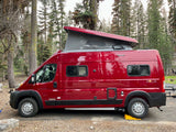 Red Winnebago Solis RV rental with pop-top raised, parked in a forest campsite, leveled with a yellow block.
