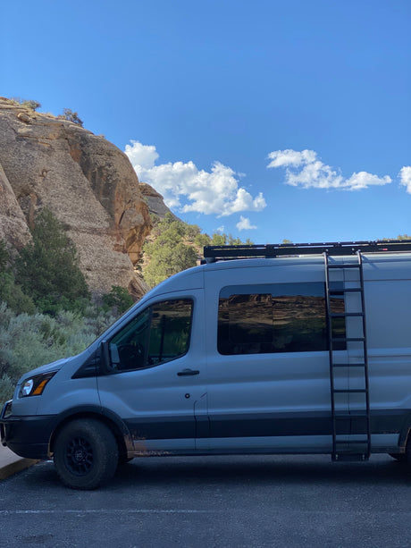 A grey camper van (an RV rental) with a ladder and roof rack is parked near a massive rocky cliff under a blue, cloudy sky.