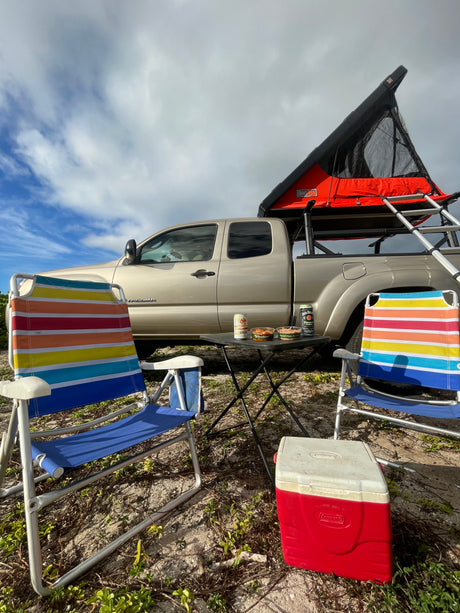 A tan truck with a rooftop tent, camp chairs, cooler, and table. An outdoor adventure awaits! Consider this an RV rental alternative.