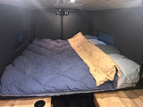 Comfortable RV rental bed setup, with blue comforter, pillows, and a tan sleeping bag in a cozy interior.