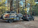Dark gray off-road camper trailer, similar to an RV rental, hitched to a truck with dirt bikes, parked in a pine forest.