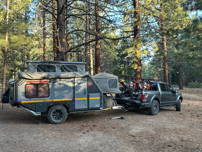 Dark gray off-road camper trailer, similar to an RV rental, hitched to a truck with dirt bikes, parked in a pine forest. — photo 19