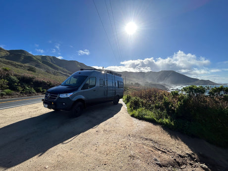This grey RV rental is perfectly parked on a scenic coastal pull-off, overlooking the ocean and rolling mountains on a bright day.
