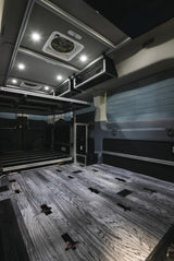 Modern RV rental interior: wood-look floor, elevated bed platform, overhead storage, and bright LED lighting.