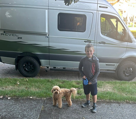 A young boy holds his curly-haired dog's leash beside a silver Winnebago Revel 4x4 rv rental, ready for adventure.