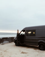 Dark grey camper van for RV rental, parked on a sandy beach with the ocean at sunset. Driver's door open for van life travel.