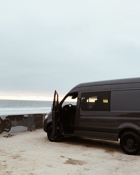 Dark grey camper van for RV rental, parked on a sandy beach with the ocean at sunset. Driver's door open for van life travel.