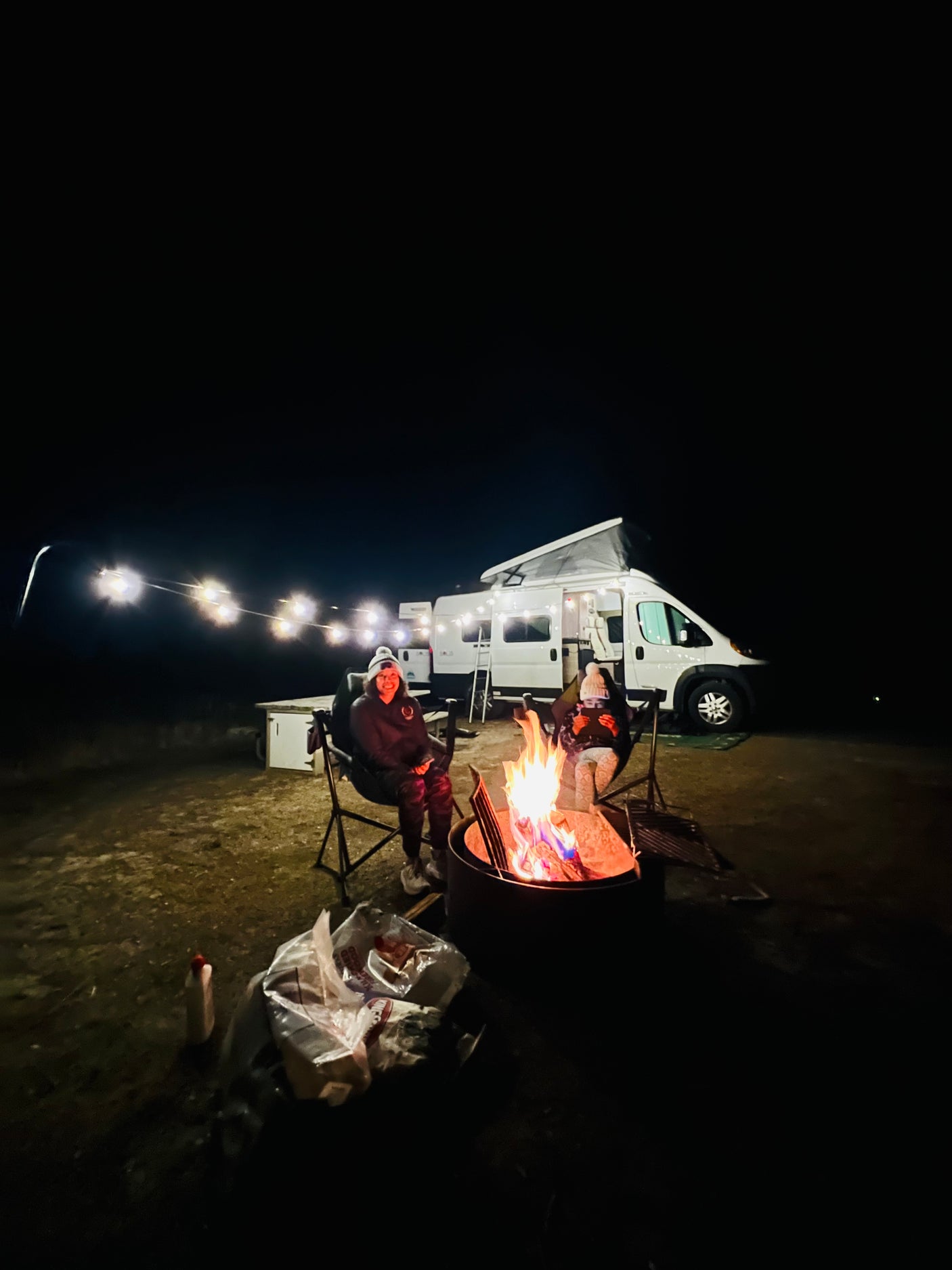 Two people enjoy a cozy campfire with an RV rental and string lights under the night sky.