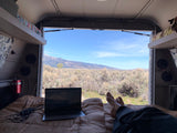 Relaxing in an RV rental, feet up on the bed with a laptop, enjoying panoramic desert mountain views from the open camper.