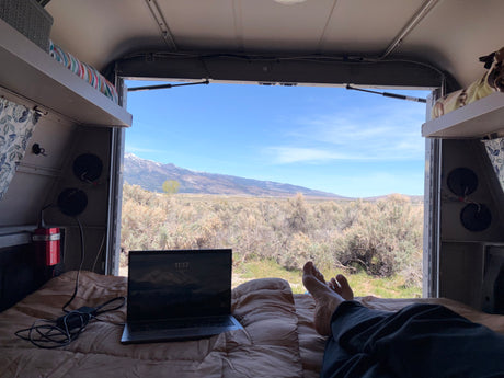 Relaxing in an RV rental, feet up on the bed with a laptop, enjoying panoramic desert mountain views from the open camper.