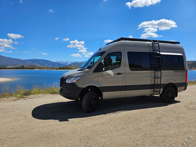 A light tan rv rental van, equipped for adventure, parked on a dirt road beside a blue lake with scenic mountains.
