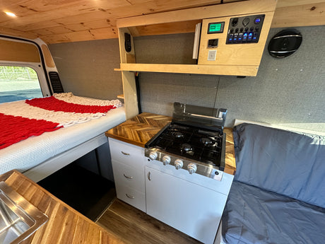 RV rental interior: This camper van features a bed, kitchen with stove and sink, wooden counters, and a control panel.