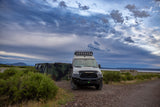 Rugged Sprinter van, ideal as an RV rental, on a dirt path near camouflage containers under a dramatic cloudy sky.