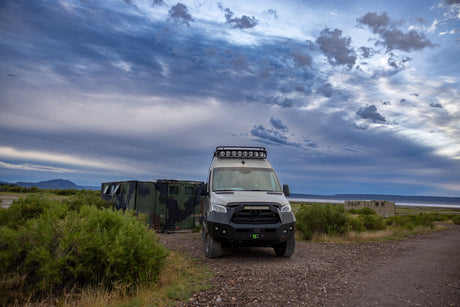 Rugged Sprinter van, ideal as an RV rental, on a dirt path near camouflage containers under a dramatic cloudy sky.