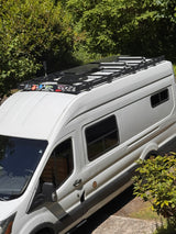 A white camper van, ideal for RV rental, showcasing rooftop solar panels, a sturdy roof rack, and unique decals.