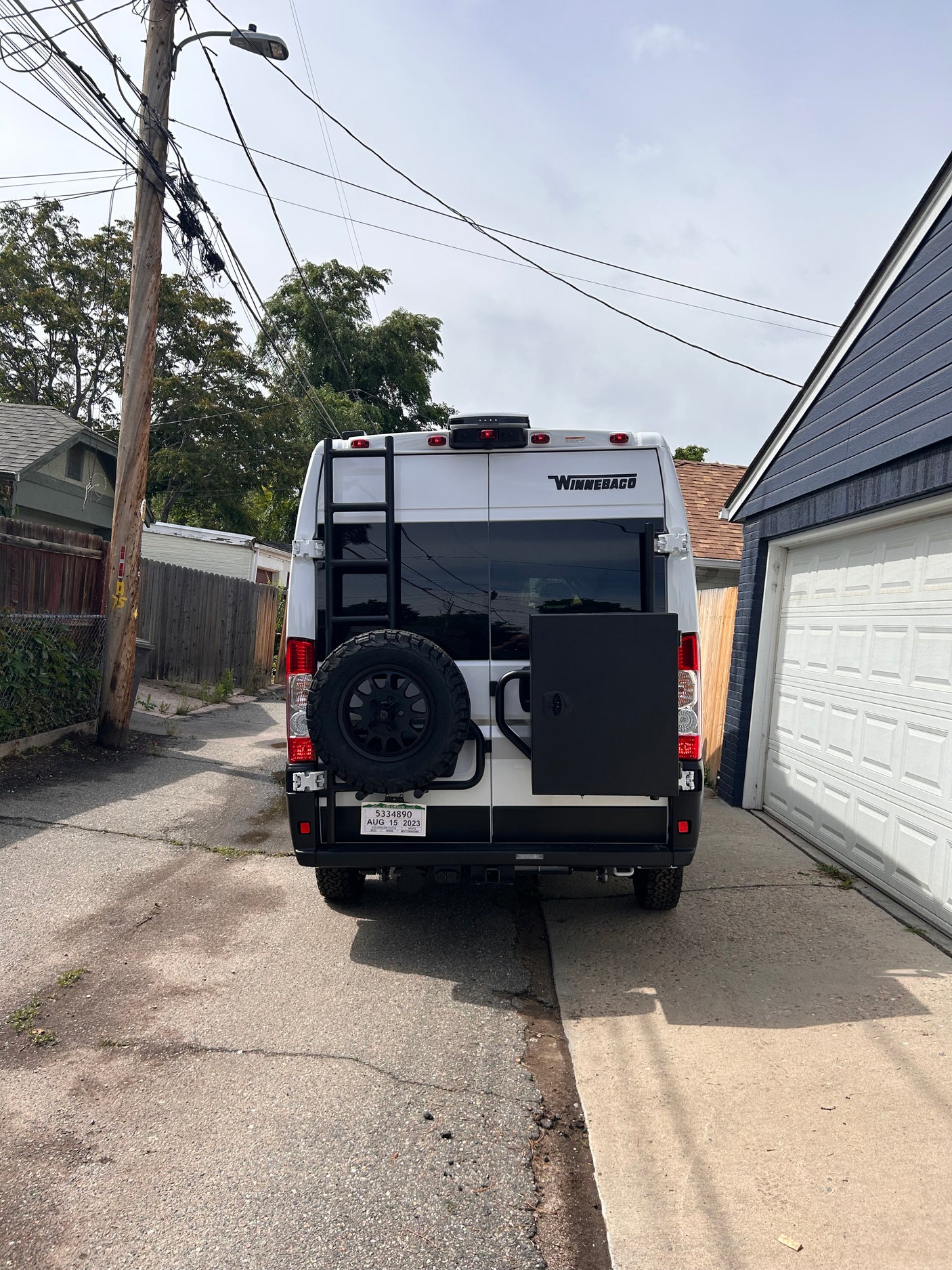Rear view of a white Winnebago rv rental camper van with a ladder, spare tire, and black storage box, parked in an alley.