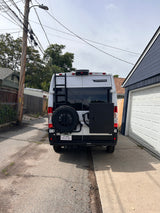 Rear view of a white Winnebago rv rental camper van with a ladder, spare tire, and black storage box, parked in an alley.