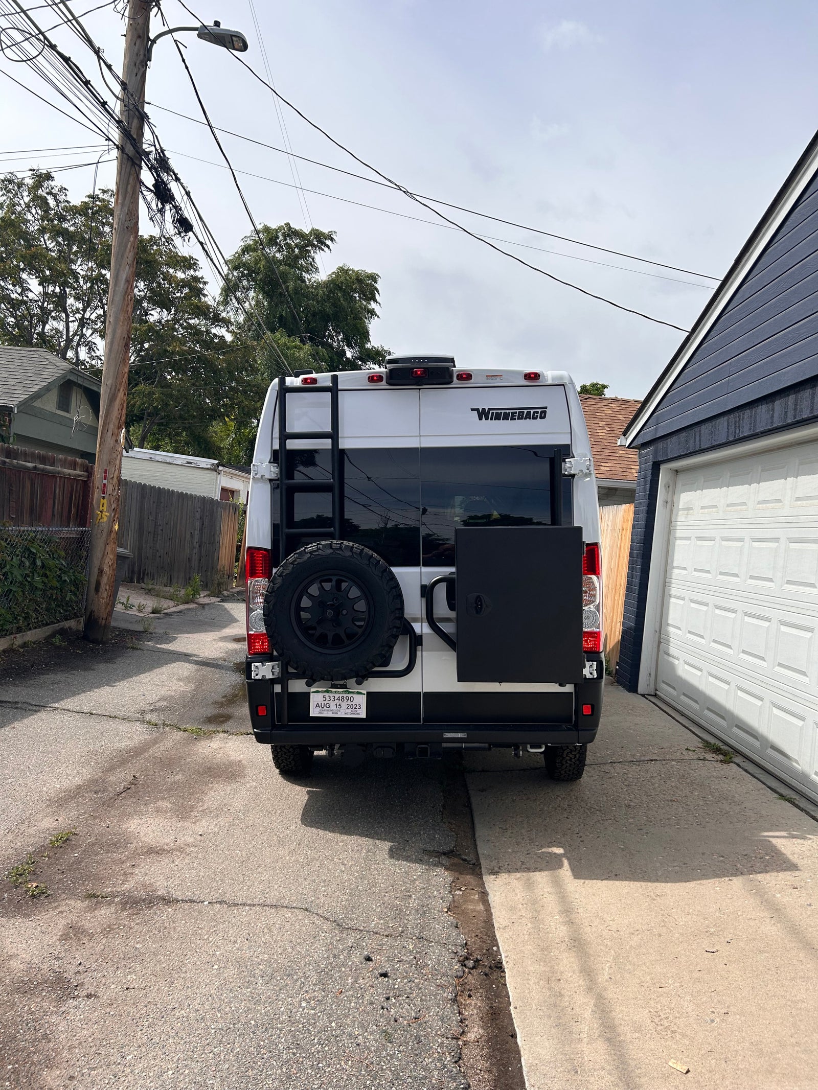 Rear view of a white Winnebago rv rental camper van with a ladder, spare tire, and black storage box, parked in an alley.