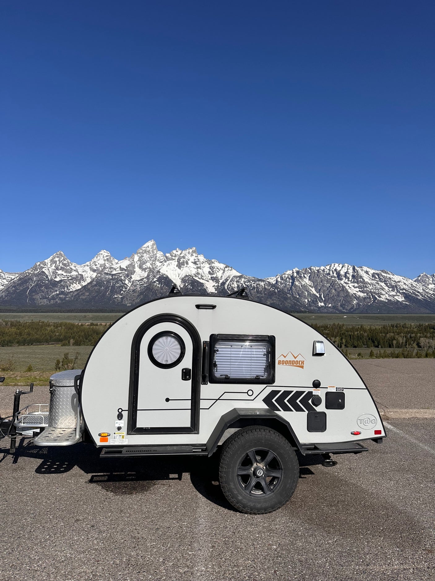 This RV rental, a sleek teardrop trailer, sits before snow-capped mountains under a clear blue sky.