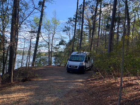 White RV rental van at a peaceful lakeside campsite, nestled among towering pine trees.