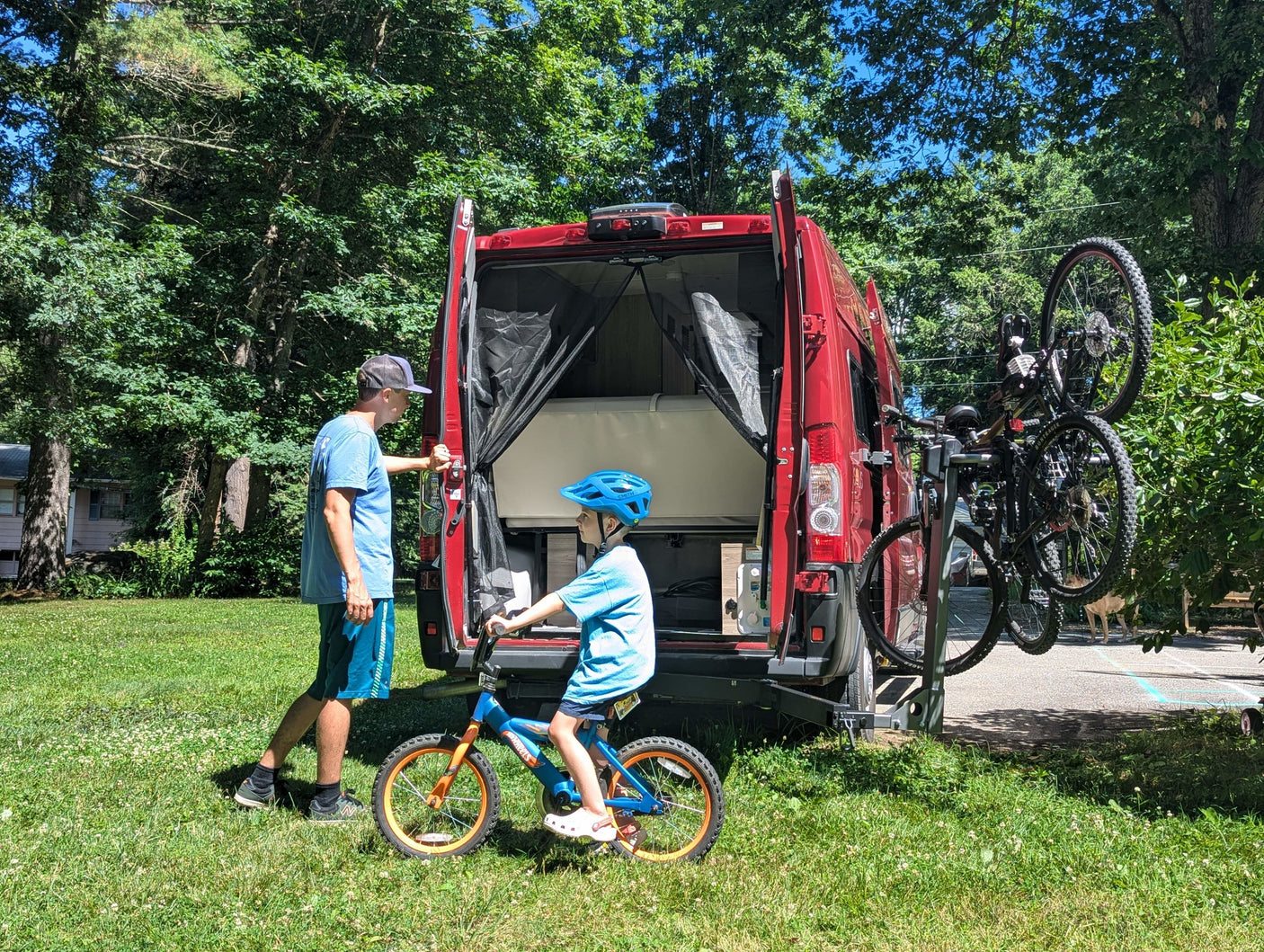 Dad & son with their **rv rental** van, bikes ready for adventure. Open rear doors show storage, while a bike rack carries two adult bikes. Child on bike with helmet.