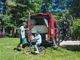 Dad & son with their **rv rental** van, bikes ready for adventure. Open rear doors show storage, while a bike rack carries two adult bikes. Child on bike with helmet.