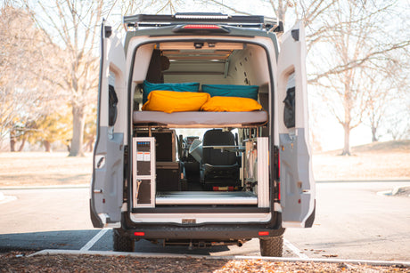 Open rear of an RV rental camper van reveals a raised bed, control panel, cooler, and passenger seat, perfect for road trips.