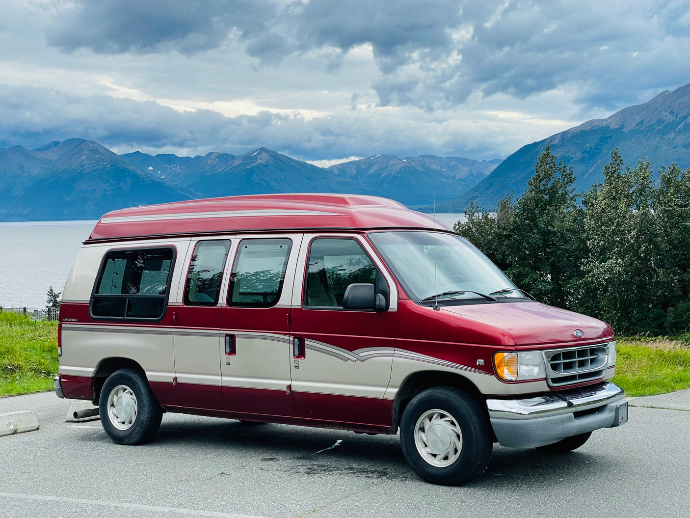 Red and white RV rental van overlooks a scenic lake and mountains under cloudy skies. Perfect for road trips!