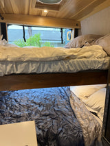Inside an RV rental, a cozy bunk bed setup. The top bunk has white bedding, and the lower bunk features a shiny gray sleeping bag.