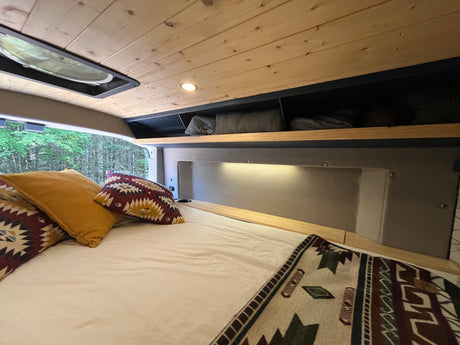 This inviting rv rental camper van interior features a comfy bed, wood ceiling, ample storage, and a picturesque forest view.