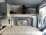Interior of an RV rental showing a textured bed, compact sink area, and overhead storage with a Luno bag. Ideal for travel.