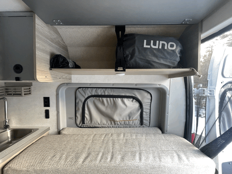 Interior of an RV rental showing a textured bed, compact sink area, and overhead storage with a Luno bag. Ideal for travel.