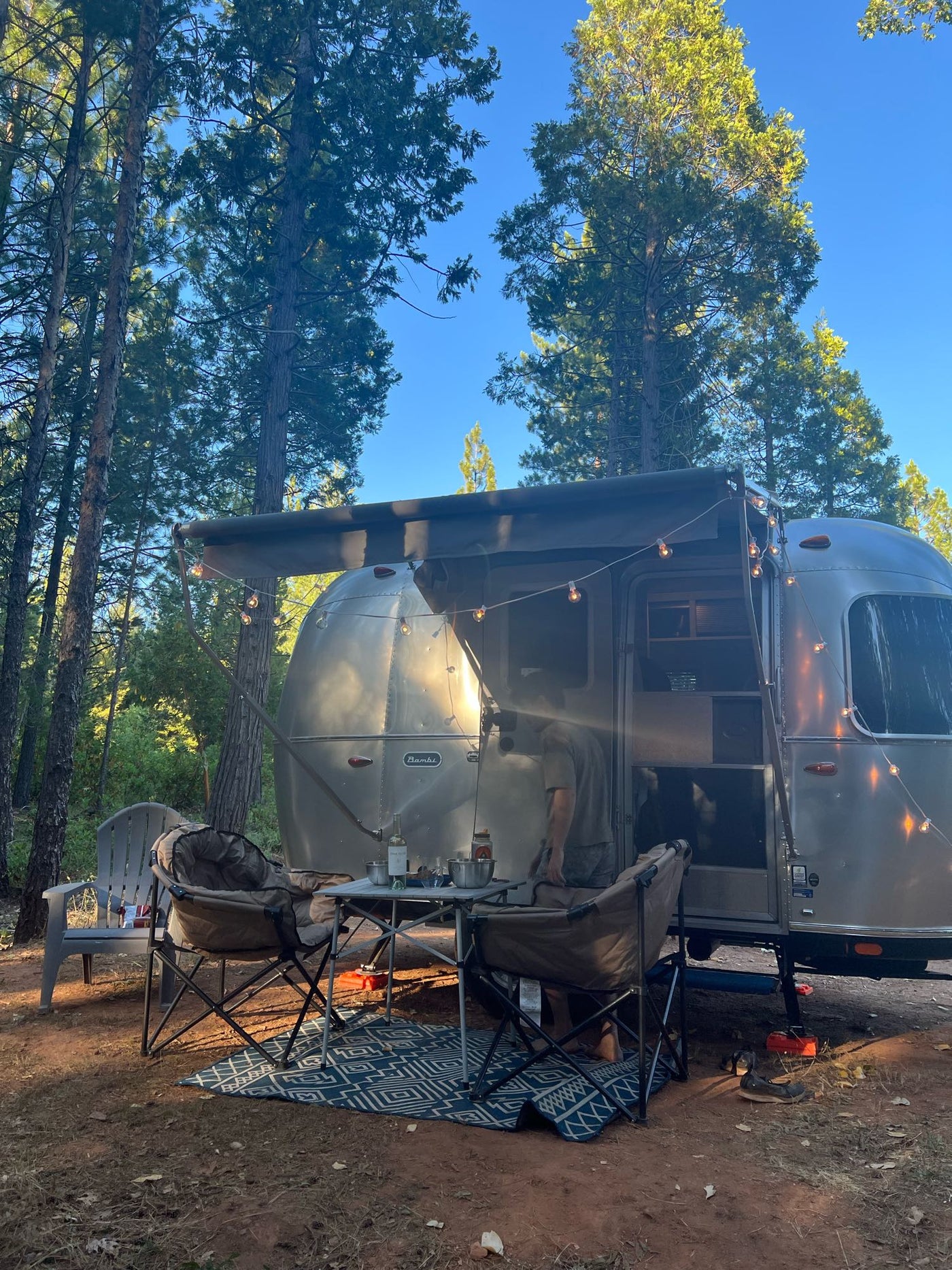  parked on rocky ground with dark trees and a blue sky.|Cozy Airstream RV rental campsite nestled in the forest with outdoor setup and string lights — photo 9