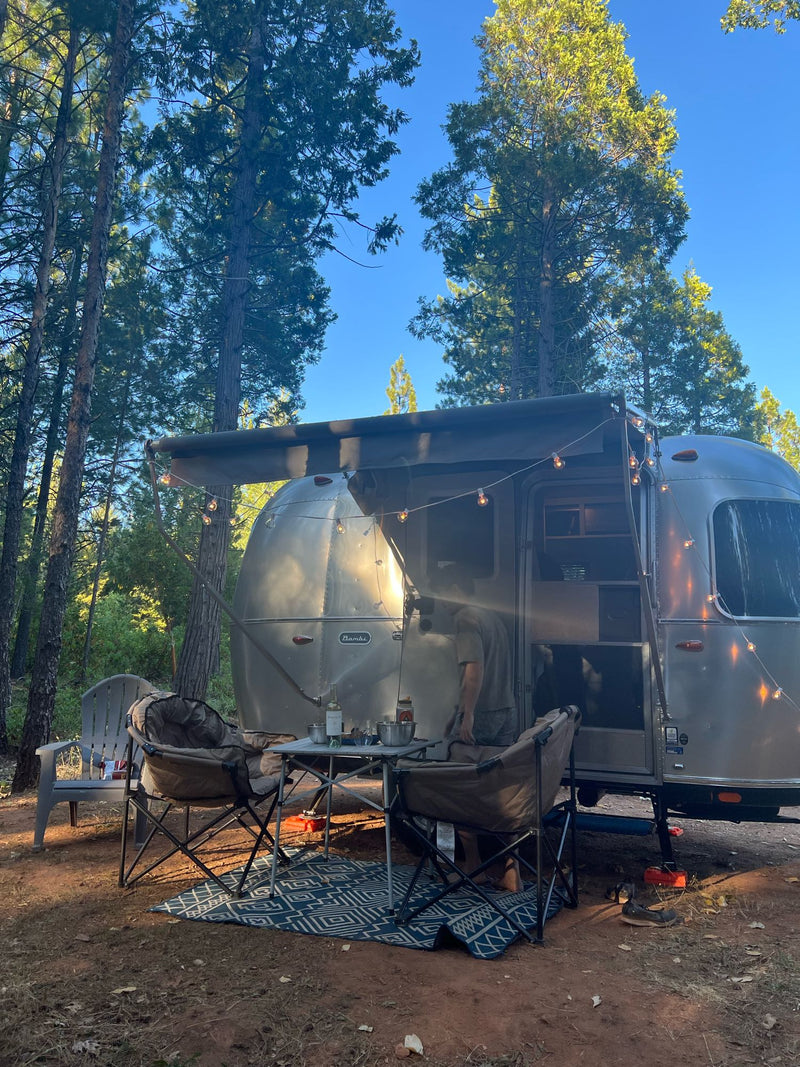  parked on rocky ground with dark trees and a blue sky.|Cozy Airstream RV rental campsite nestled in the forest with outdoor setup and string lights — photo 9