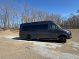 Dark grey Sprinter van RV rental on a dirt path with bare trees and distant volleyball nets. Ideal for adventure travel.