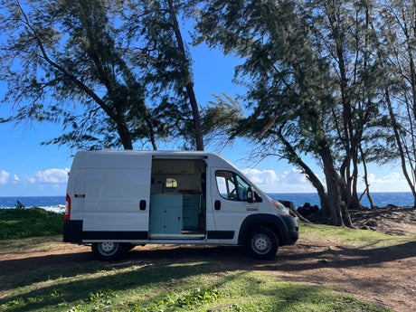 White RV rental van parked by the ocean under tall trees. Its open side reveals a customized interior with light blue cabinets. Ideal for van life.