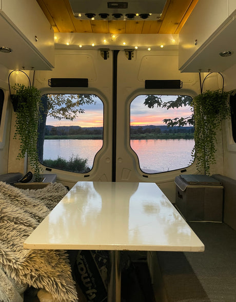Inside a spacious RV rental, watching a vibrant sunset over a river from the comfortable, lamp-lit dining area.