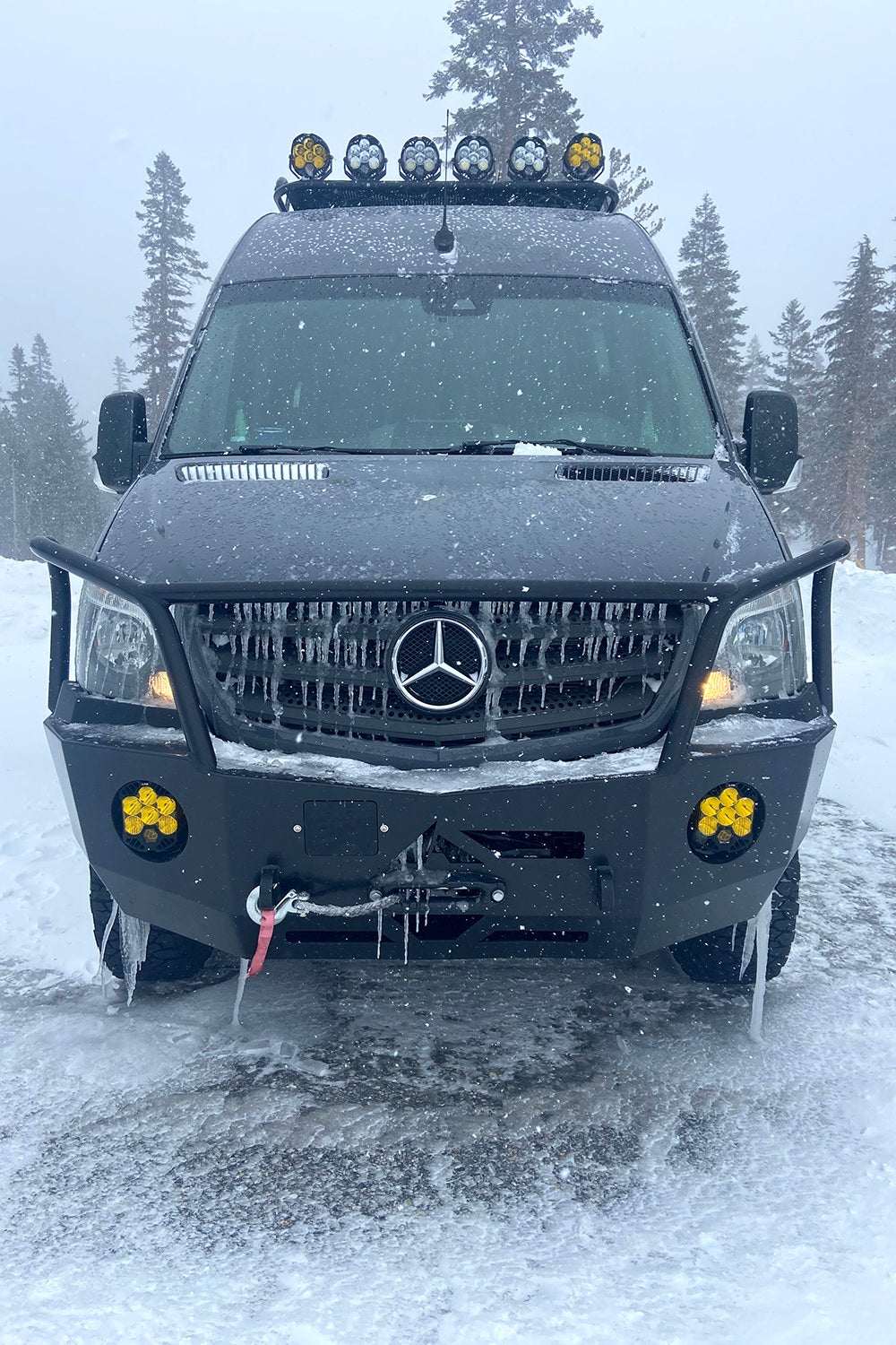 Front view of a black Mercedes Sprinter RV rental, covered in snow and icicles, ready for rugged winter adventures.